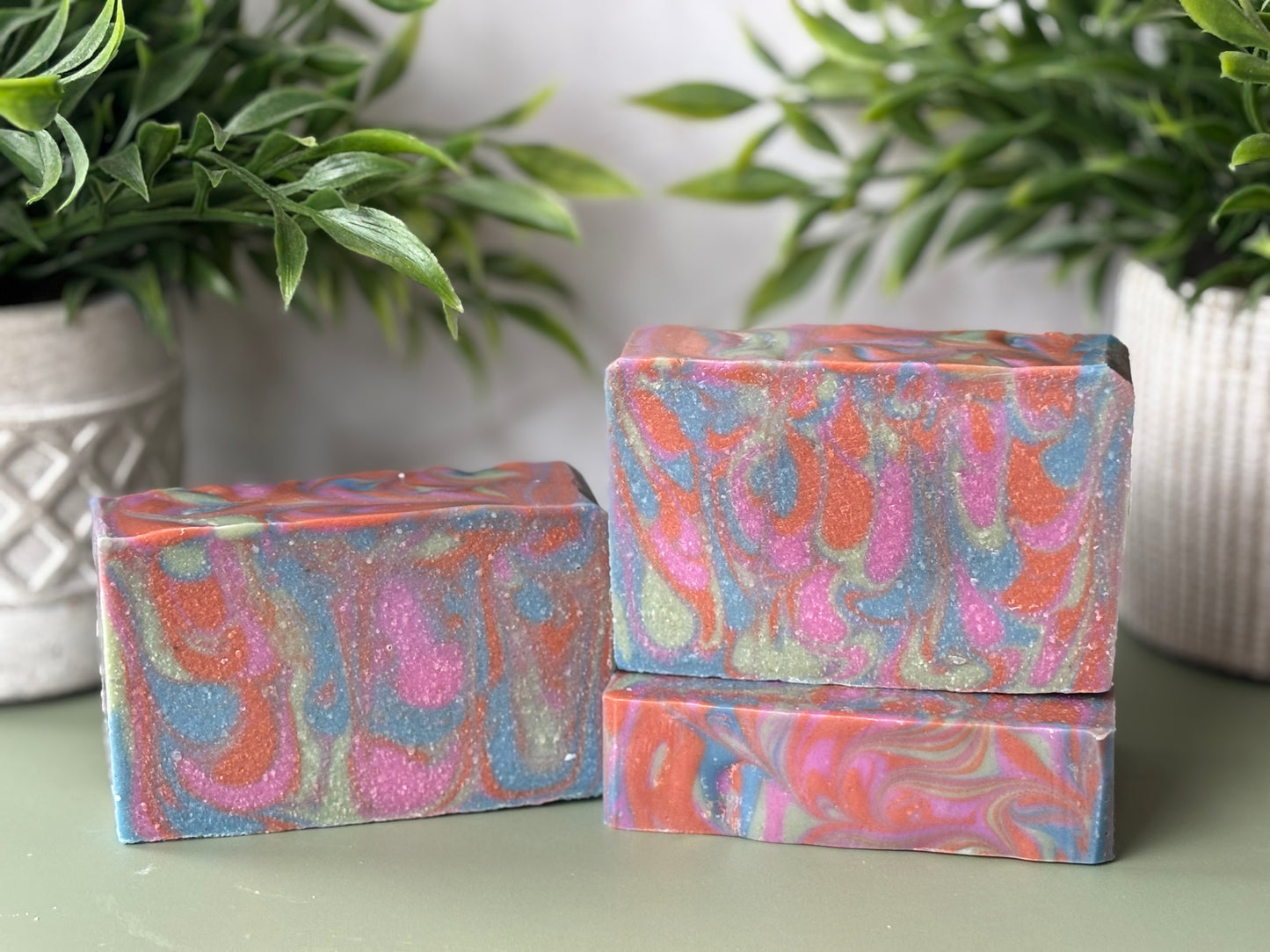 Three colorful soap bars with a marbled pattern on a light surface, with plants in the background.