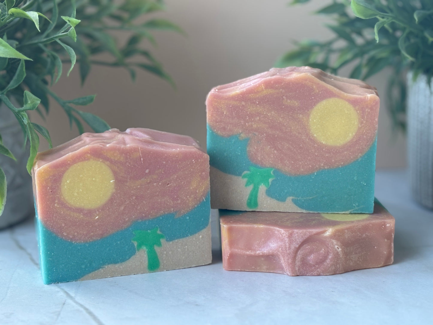 Three bars of soap with artistic designs resembling a beach landscape on a light surface with plants in the background.
