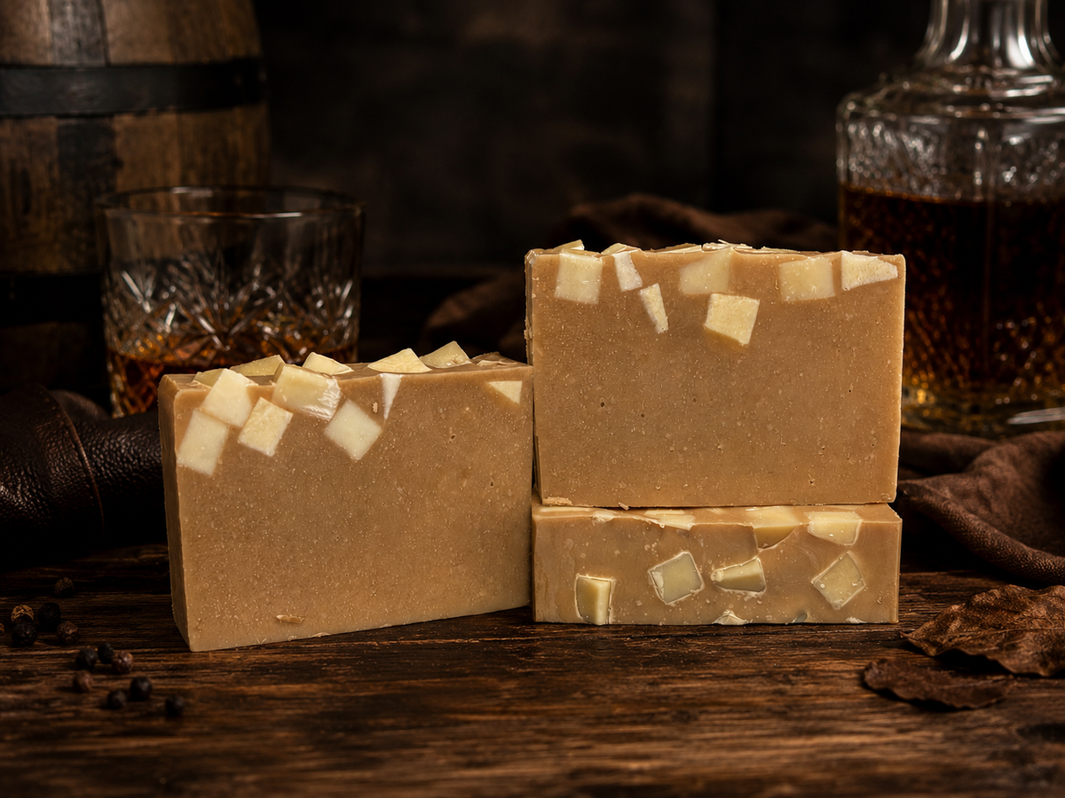 Three rectangular bars of handmade soap in warm tan and cream tones, decorated with small pale square inclusions, displayed on a rustic wooden surface with a whiskey glass, bottle, and dark cloth in the background.