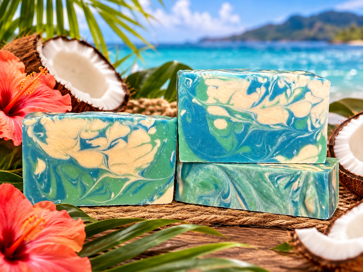 Three marbled soap bars in blue, green, and cream are arranged on a rope with tropical props including hibiscus flowers, palm leaves, and coconut halves, with a beach and ocean in the background.