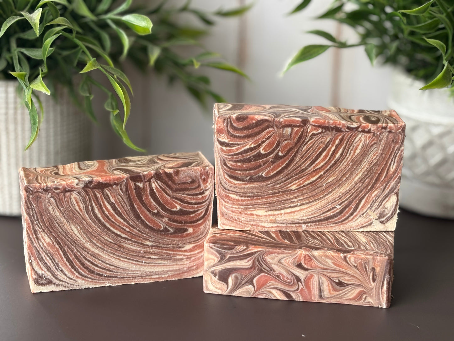 Three marbled soap bars on a dark surface with green plants in the background