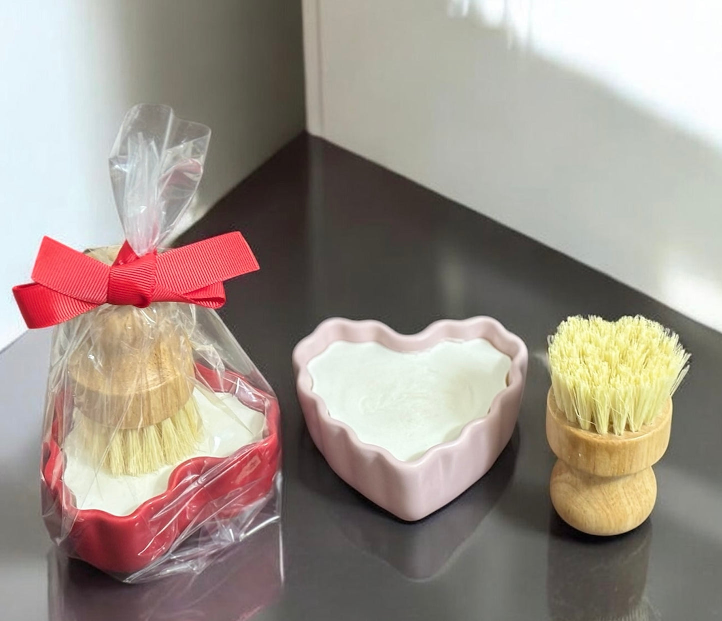 Set of kitchen cleaning brush and a heart-shaped ramekin of dish soap on a dark surface.