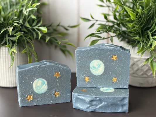 Three starry night-themed soap bars on a dark surface with plants in the background