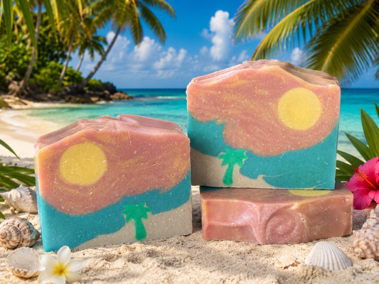Three colorful soap bars with tropical designs on a sandy beach with palm trees and ocean in the background.