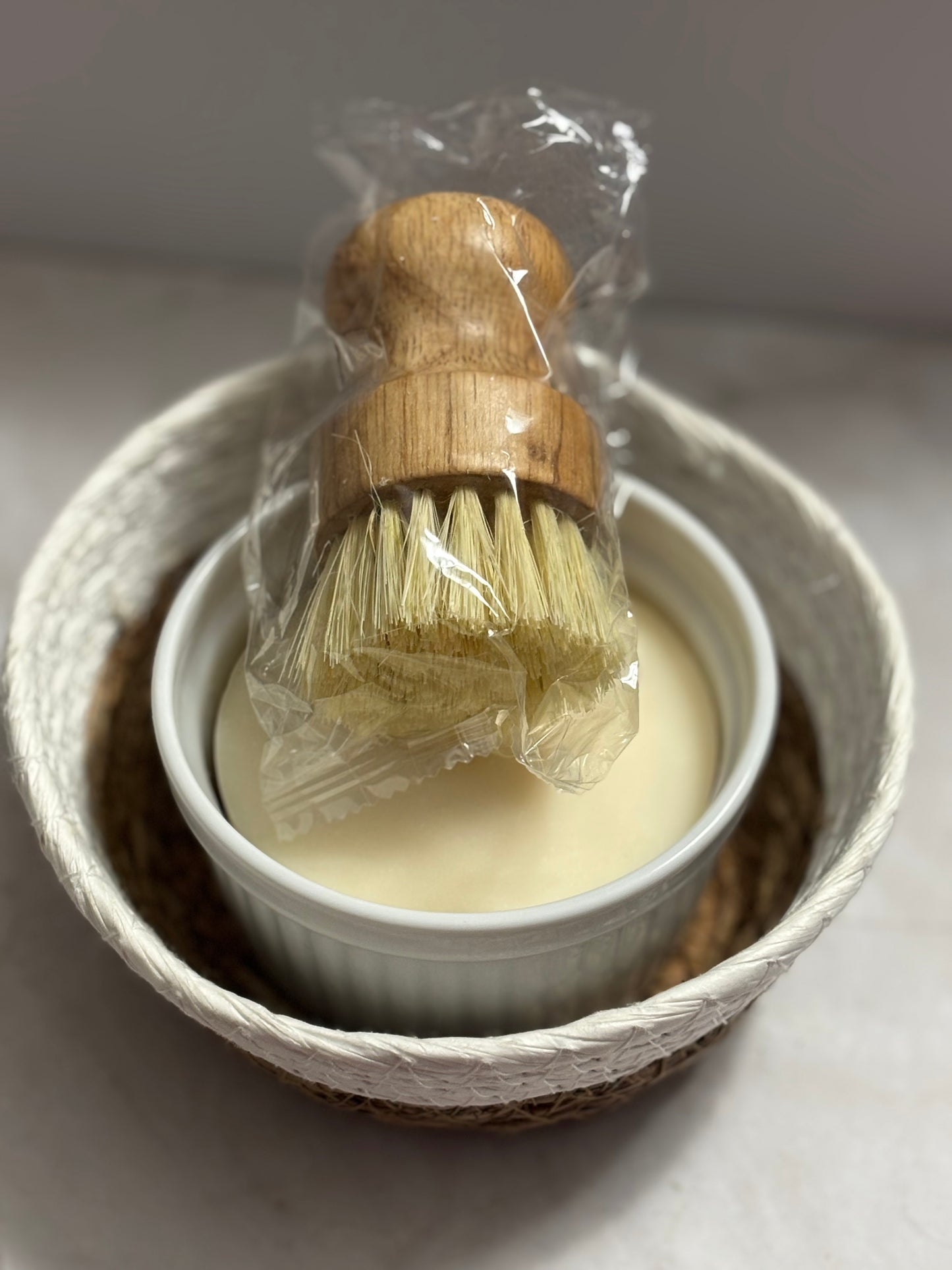 Wooden brush with bristles in a small white container of solid dish soap in a basket on a light gray background