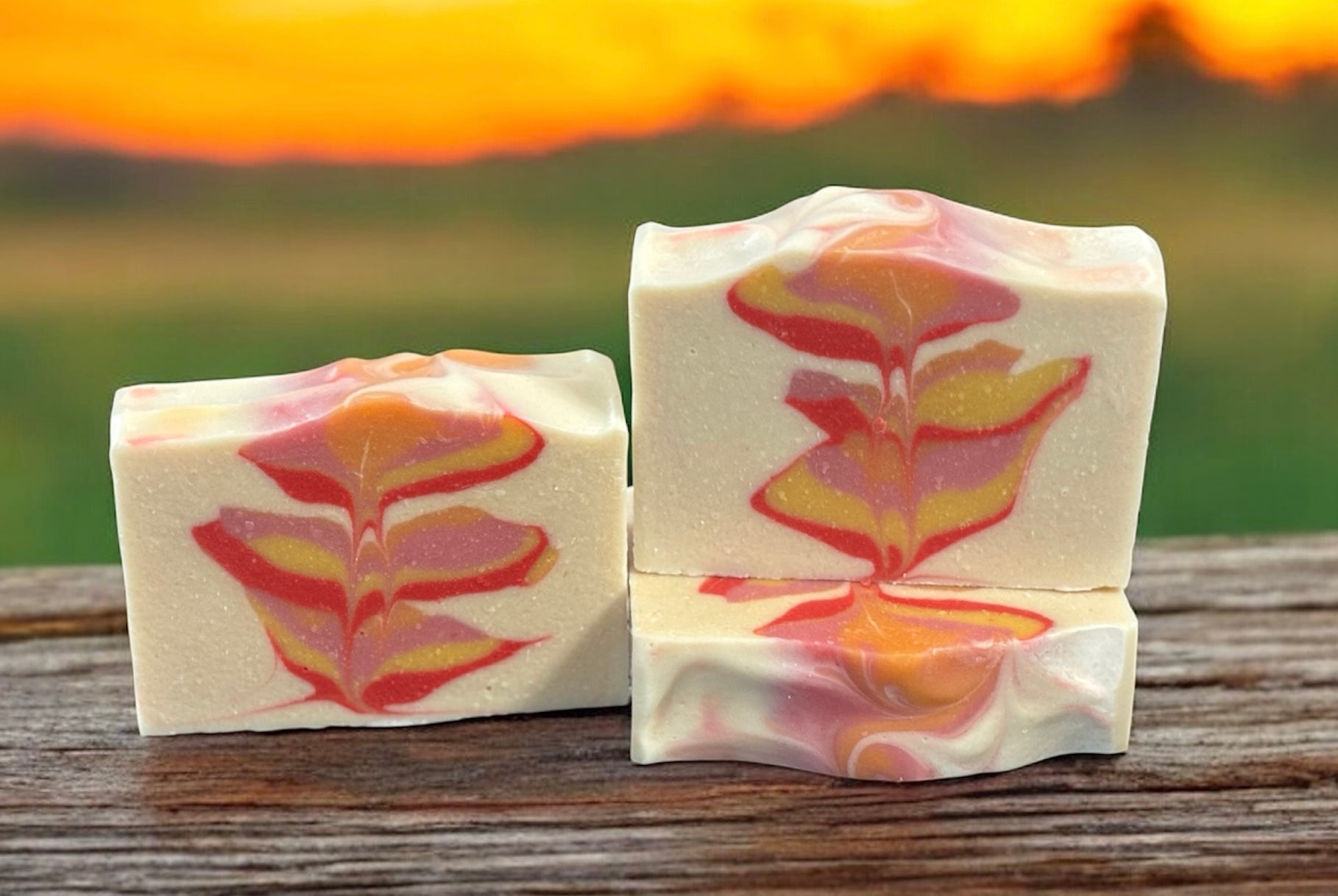 Three bars of soap with an orange, yellow, pink and red leaf pattern on a wooden surface with a sunset background