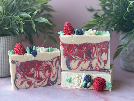 Three bars of soap with a marbled design, decorated with strawberries, blueberries, blackberries and flowers on a light surface with plants in the background.