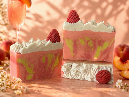 Three pink soap bars with white and red decorations on a wooden surface with peaches and flowers.