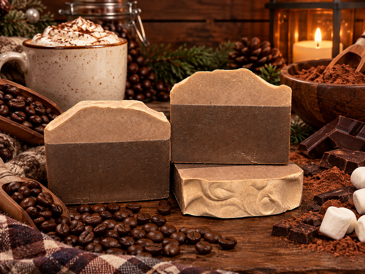 Three artisanal bars of soap in earthy brown and beige tones, styled on a rustic wooden table with coffee beans, cocoa powder, chocolate pieces, a mug of hot cocoa with whipped cream, pinecones, and a warm candle in the background.