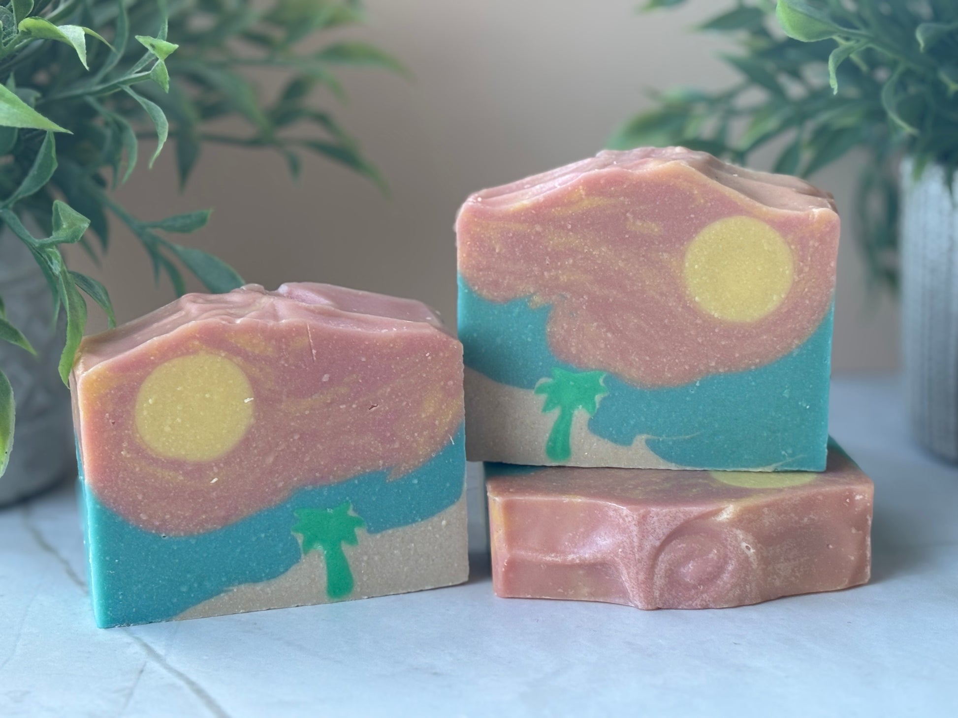 Three bars of soap with artistic designs resembling a beach landscape on a light surface with plants in the background.