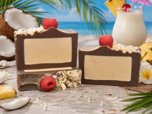 Three rectangular bars of brown soap with cream-colored centers, decorated with red cherries and shredded coconut, displayed on a tropical beach-themed surface with coconuts, pineapple, and a tiki drink in the background.