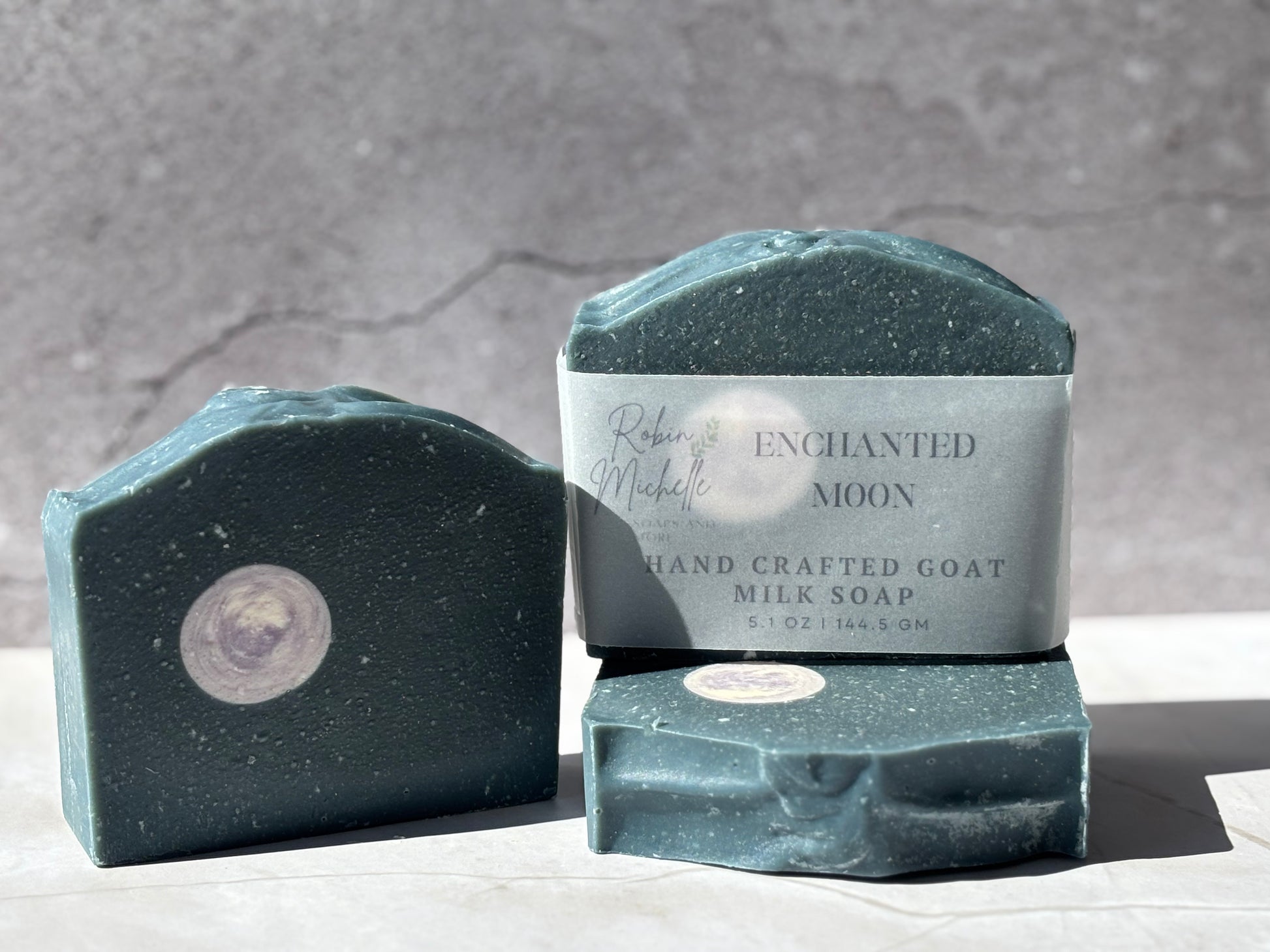 Handcrafted goat milk soap with 'Enchanted Moon' branding on a gray background. It’s a dark blue soap with shredded white soap for “stars” and a purple and white, round, moon shaped embed.