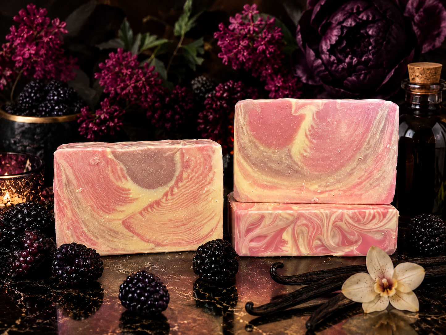 Three rectangular artisan soap bars with pink, purple and cream marbled swirls are displayed on a dark reflective surface, surrounded by blackberries, vanilla beans, a white orchid flower, and a dark bottle, with dark purple flowers in the background.