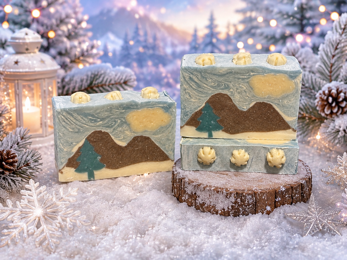Three decorative soap bars with a winter mountain landscape design, displayed on a snowy surface with a rustic wood slice. The background is a festive, blurred Christmas forest scene with lights, pine trees, and a lantern.