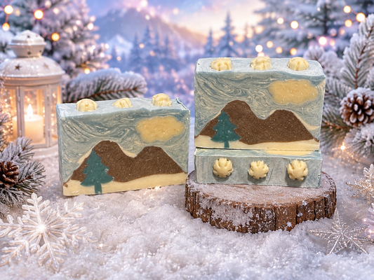 Three decorative soap bars with a winter mountain landscape design, displayed on a snowy surface with a rustic wood slice. The background is a festive, blurred Christmas forest scene with lights, pine trees, and a lantern.