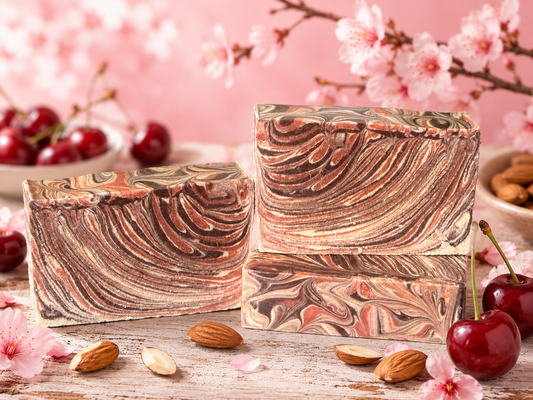 Three rectangular bars of marbled soap with brown, beige, and pink swirls, arranged on a rustic wooden surface with cherries, almonds, and pink blossoms in the background.
