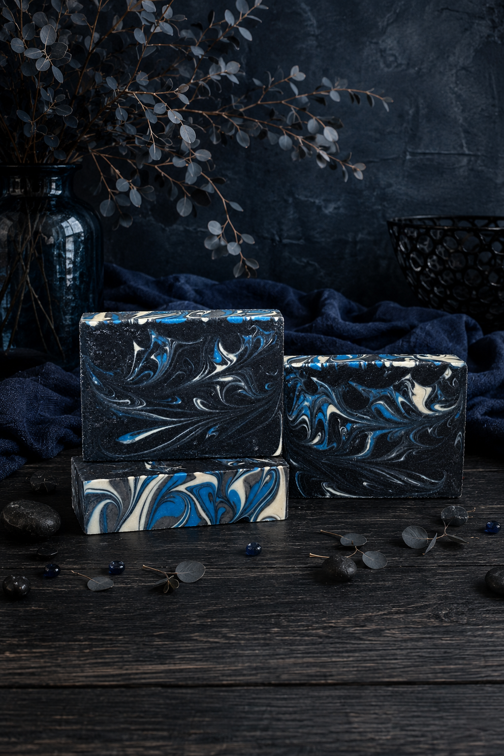 Three rectangular bars of soap with a marbled swirl pattern in black, blue, and white, arranged on a dark wooden surface with a dark blue cloth, dried leaves, and a dark textured background.