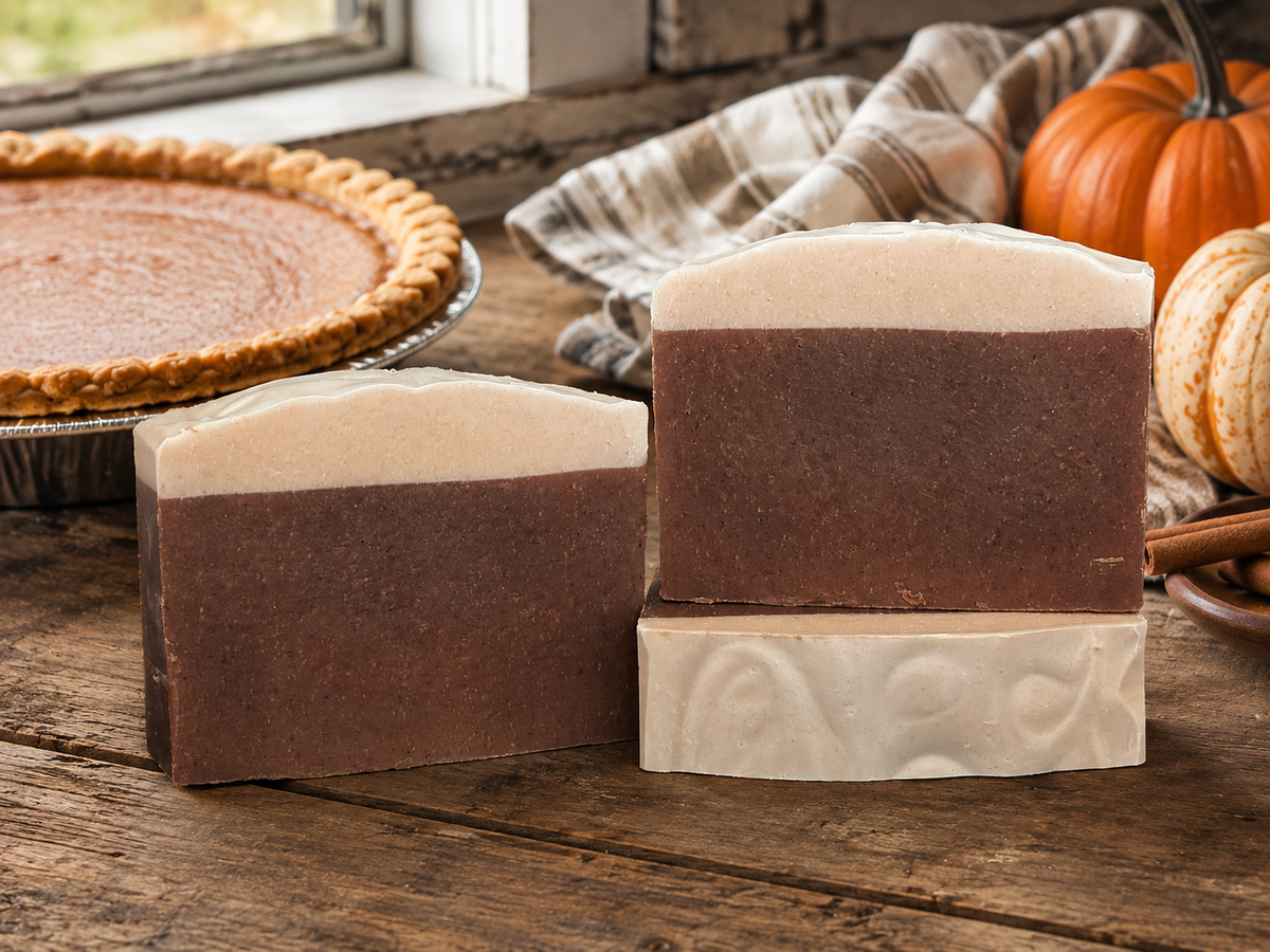 Three handmade soap bars with a dark brown and cream color-blocked design, styled on a rustic wooden table next to a pumpkin pie and autumn pumpkins.