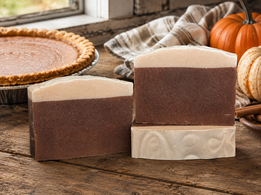 Three handmade soap bars with a dark brown and cream color-blocked design, styled on a rustic wooden table next to a pumpkin pie and autumn pumpkins.
