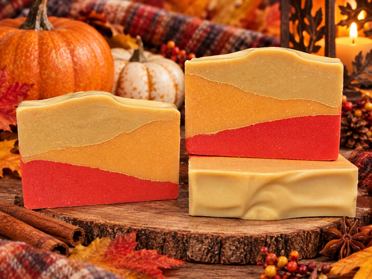Three handmade layered soap bars with a yellow, orange, and red color scheme, styled on a rustic wood slice with autumn props like pumpkins, cinnamon sticks, and a lit candle.